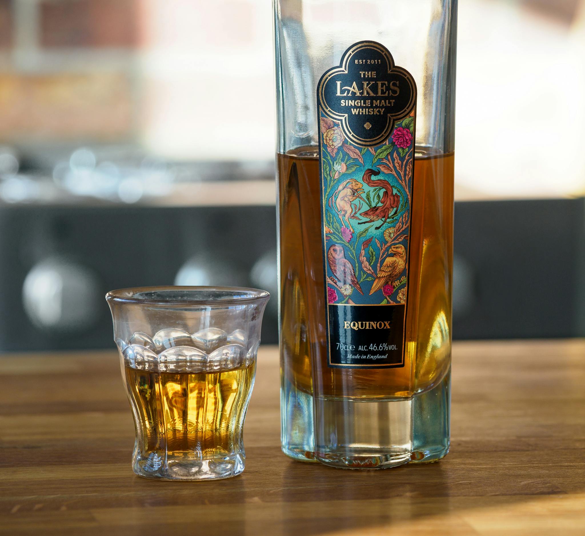 A close-up of The Lakes single malt whisky bottle and glass on a wooden table.